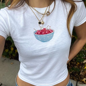 以下が含まれることがあります： 赤いチェリーがたっぷり入った青いボウルが描かれた白いTシャツ。Tシャツはクロップド丈で、ゆったりとしたフィット感です。