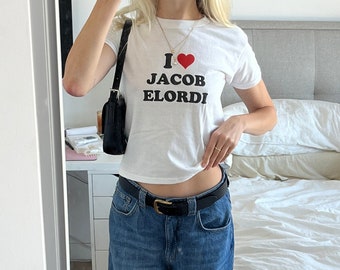 Op de afbeelding: Een vrouw draagt een wit, gecropped T-shirt met de tekst "I love Jacob Elordi" en een rood hart. Ze draagt ook een blauwe jeans en een zwarte riem.