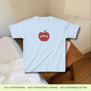 Pu&ograve; includere: Maglietta blu chiaro con un grafico di una mela rossa e il nome "Fiona" scritto su di essa.