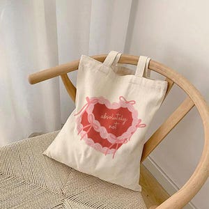 Pode incluir: Uma bolsa tote de lona branca com um design em forma de coração rosa e vermelho. O coração tem fitas rosa e o texto "absolutely not" dentro.