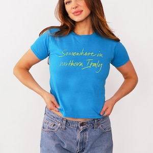 May include: A woman wearing a blue t-shirt with the text "Somewhere in northern Italy" in yellow lettering. She is wearing blue jeans.