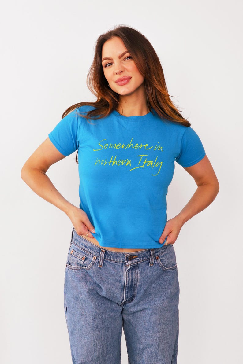 May include: A woman wearing a blue t-shirt with the text "Somewhere in northern Italy" in yellow lettering. She is wearing blue jeans.