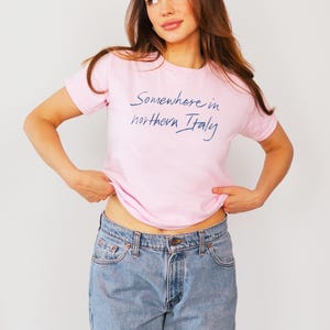 May include: A woman wearing a pink t-shirt with the text "Somewhere in northern Italy" in blue script. She is wearing blue jeans and is standing in front of a white background.