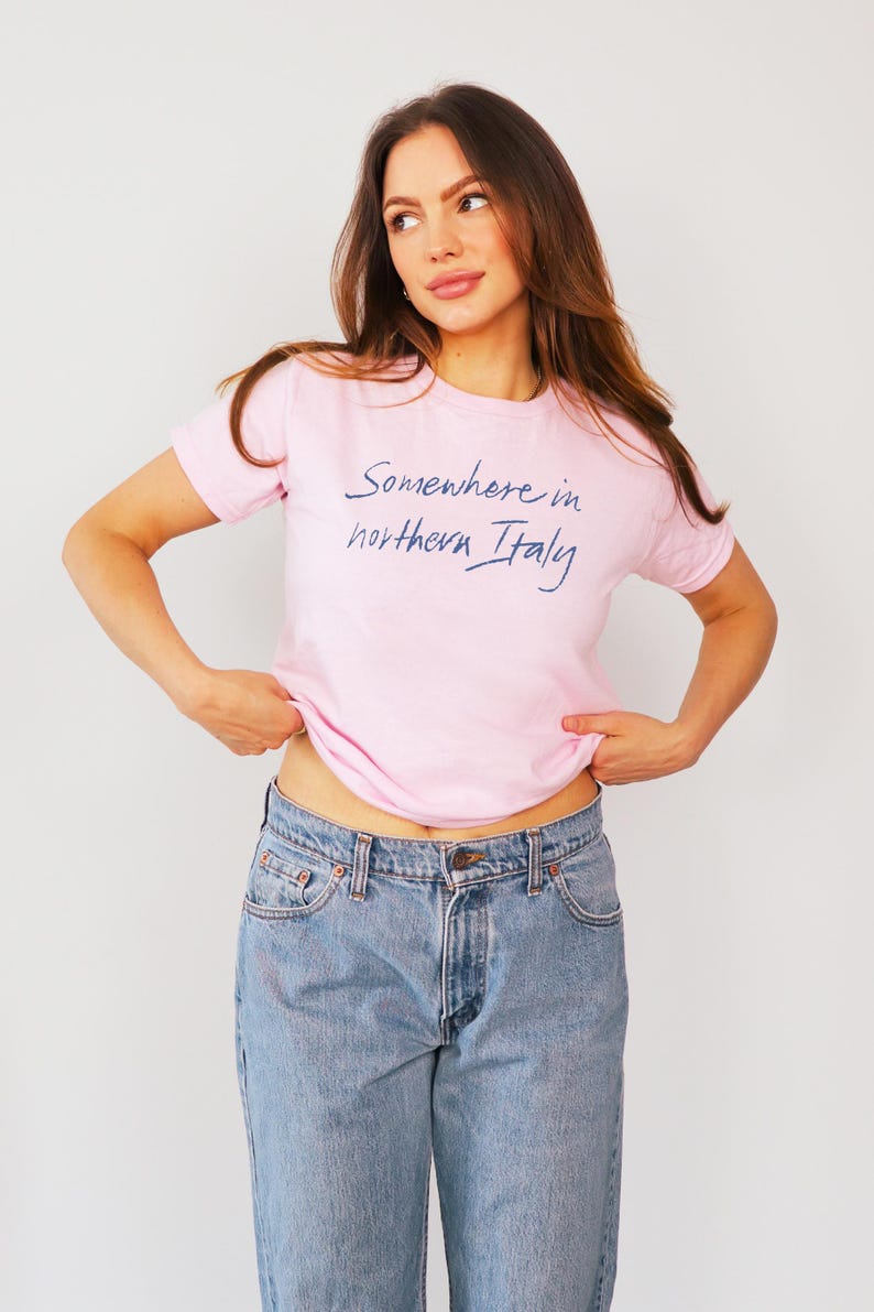 May include: A woman wearing a pink t-shirt with the text "Somewhere in northern Italy" in blue script. She is wearing blue jeans and is standing in front of a white background.