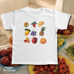 May include: White t-shirt with a colorful fruit print. The fruits include a papaya, strawberry, lemon, grapes, pineapple, grapefruit, and orange.
