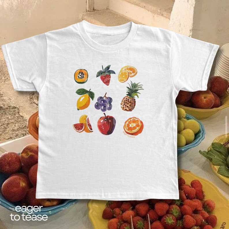 May include: White t-shirt with a colorful fruit print. The fruits include a papaya, strawberry, lemon, grapes, pineapple, grapefruit, and orange.