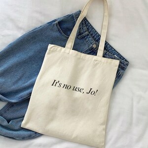 Pode incluir: Uma bolsa tote de lona bege com al&ccedil;as pretas e o texto "It's no use, Jo!" impresso na frente.