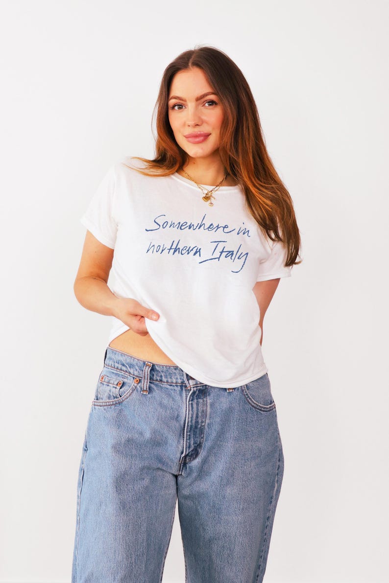 May include: A white t-shirt with a blue inscription that reads "Somewhere in northern Italy". The model is wearing blue jeans.