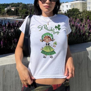 Pu&ograve; includere: Maglietta bianca con un trifoglio verde e il testo "St. Patrick's Day". La maglietta presenta un'illustrazione di un cartone animato di una ragazza con un vestito verde che tiene un trifoglio.