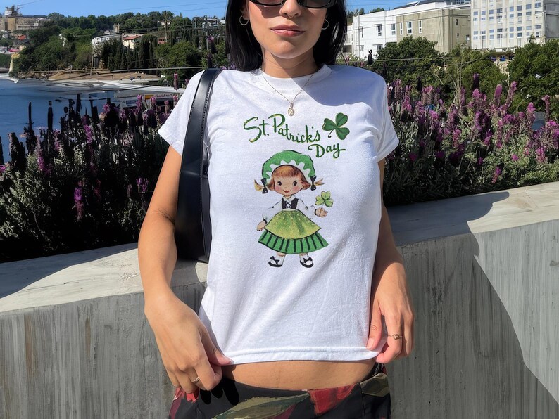 Pu&ograve; includere: Maglietta bianca con un trifoglio verde e il testo "St. Patrick's Day". La maglietta presenta un'illustrazione di un cartone animato di una ragazza con un vestito verde che tiene un trifoglio.