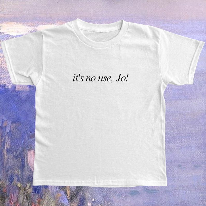 May include: White crew neck t-shirt with the phrase "it's no use, Jo!" printed in black. The shirt is made of a soft, lightweight fabric. The background is a soft purple and blue abstract painting.