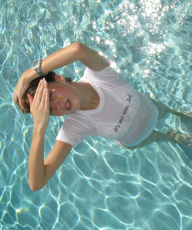 May include: A person wearing a white t-shirt with the text "it's no use, Jo!" is submerged in a pool. The water is clear and reflects sunlight. A silver watch is visible on the person's wrist.