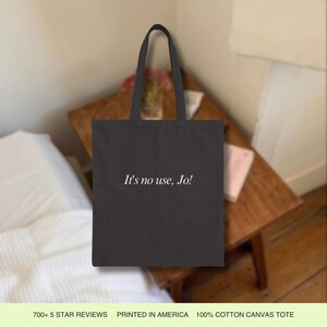 Pode incluir: Uma bolsa tote de lona preta com o texto "It's no use, Jo!" impresso em branco. A bolsa est&aacute; sobre uma mesa de madeira.