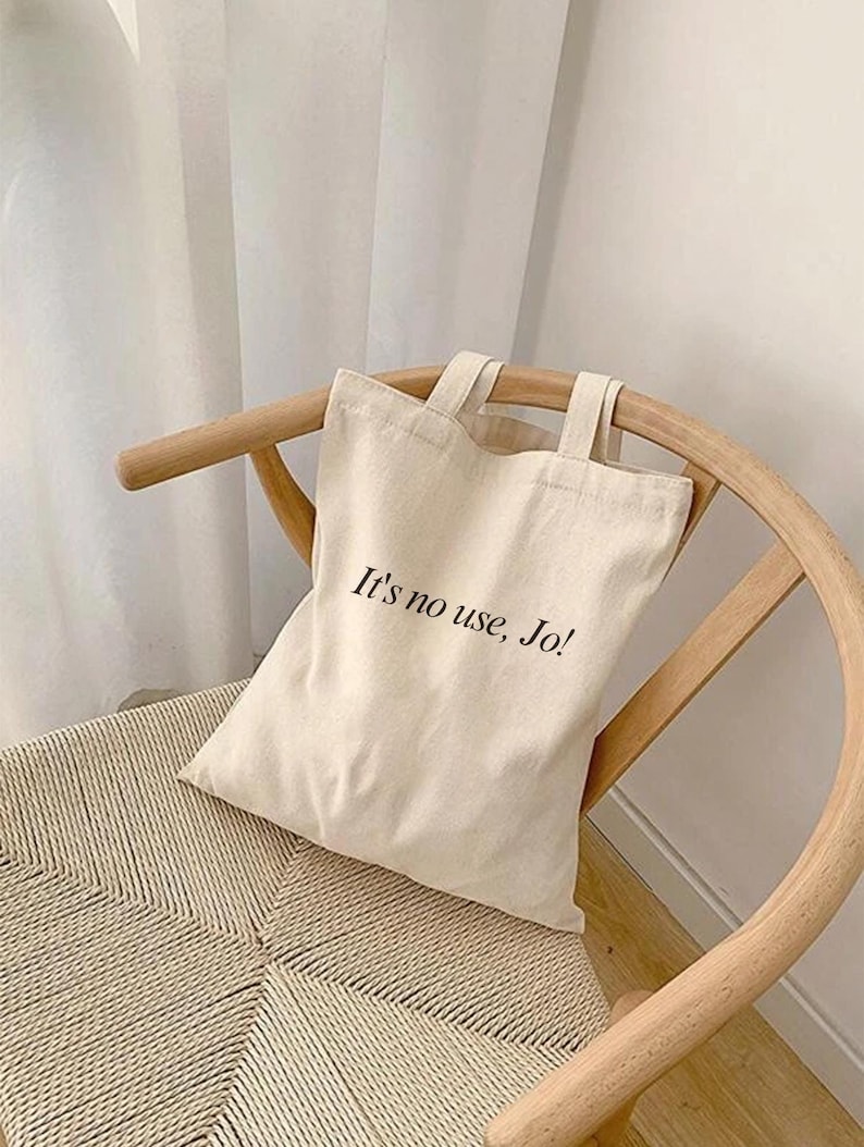Pode incluir: Uma bolsa tote de lona branca com texto preto que diz "It's no use, Jo!" A bolsa est&aacute; sentada em uma cadeira de vime marrom claro.