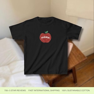 Pu&ograve; includere: Maglietta nera con una grafica di una mela rossa e il nome "Fiona" scritto su di essa.