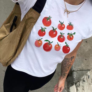 Puede incluir: Camiseta blanca con un gráfico de tomates rojos. La camiseta tiene una bolsa de bandolera marrón colgada del hombro.