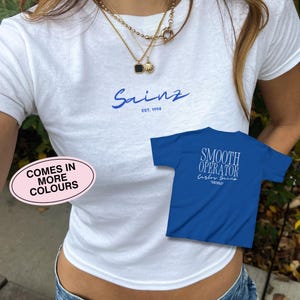 以下が含まれることがあります： 白いTシャツに青い文字で「Sainz Est. 1994」と書いてあります。白い文字で「Smooth Operator Carlos Sainz」と書いてある青いTシャツが、白いTシャツの前側に一部見えています。「Comes in More Colours」という文字がピンクの楕円形の中にあります。
