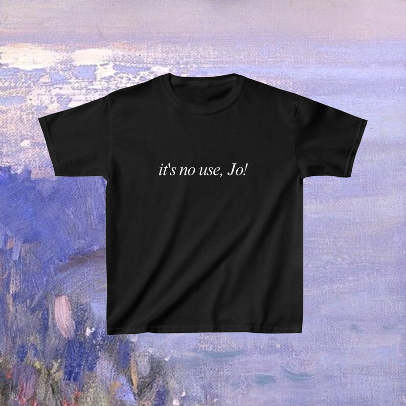 May include: Black t-shirt with the phrase "it's no use, Jo!" printed in white. The shirt is displayed against a backdrop of blue and purple brushstrokes, creating a soft, artistic background.
