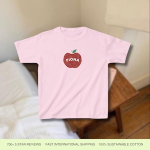 Pu&ograve; includere: Maglietta rosa con un grafico di una mela rossa e il nome "Fiona" stampato su di essa.