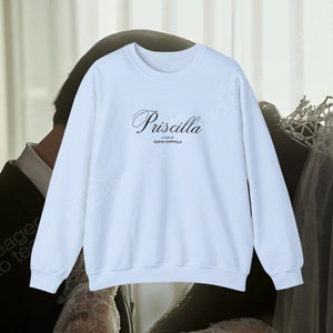 Peut inclure: Un sweat-shirt bleu clair avec le texte "Priscilla, A film by Sofia Coppola" imprimé sur le devant.