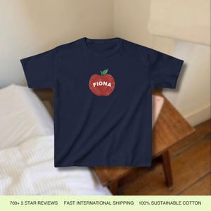 Pu&ograve; includere: Una t-shirt blu navy con una grafica di una mela rossa e il nome "Fiona" scritto su di essa.