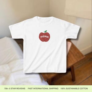 Pu&ograve; includere: Maglietta bianca con un grafico di una mela rossa e il nome "Fiona" stampato sulla mela.