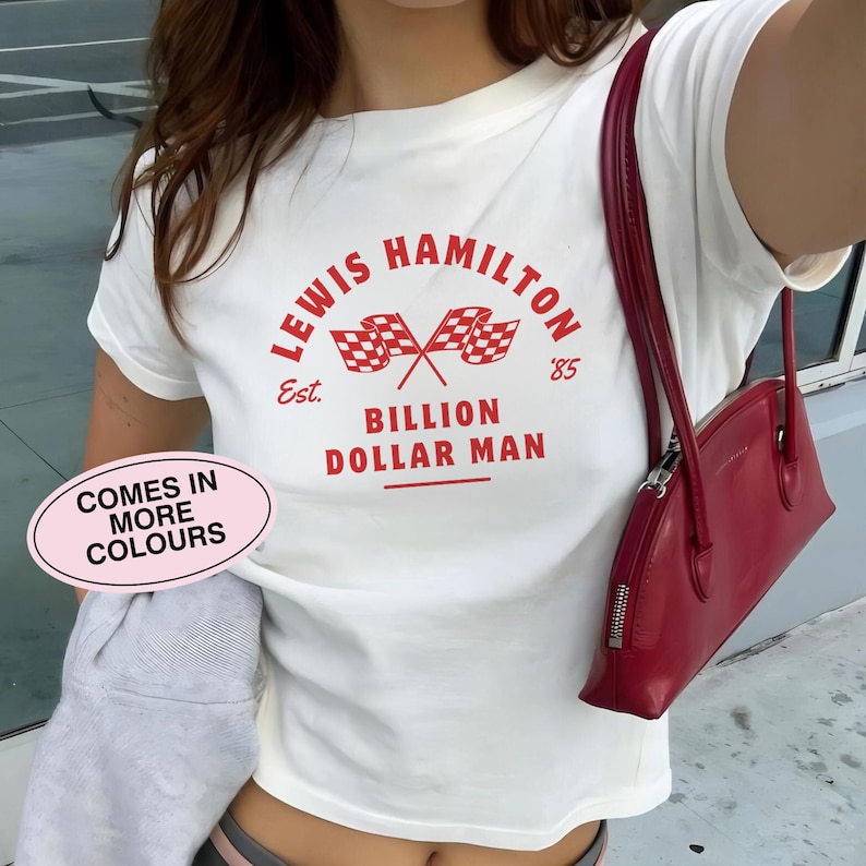 K&ouml;nnte beinhalten: Wei&szlig;es T-Shirt mit einem roten Grafikdruck, auf dem "Lewis Hamilton Est. '85 Billion Dollar Man" mit einem karierten Flaggenmuster steht.