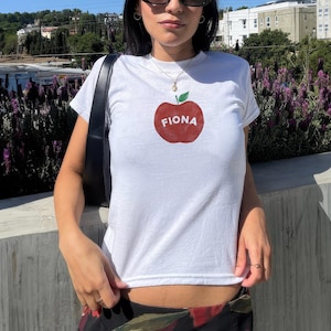 Pu&ograve; includere: Maglietta bianca con un grafico di una mela rossa e il nome "Fiona" scritto su di essa.