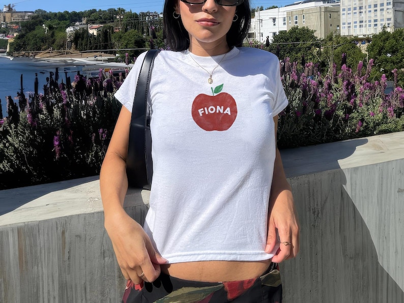Pu&ograve; includere: Maglietta bianca con un grafico di una mela rossa e il nome "Fiona" scritto su di essa.