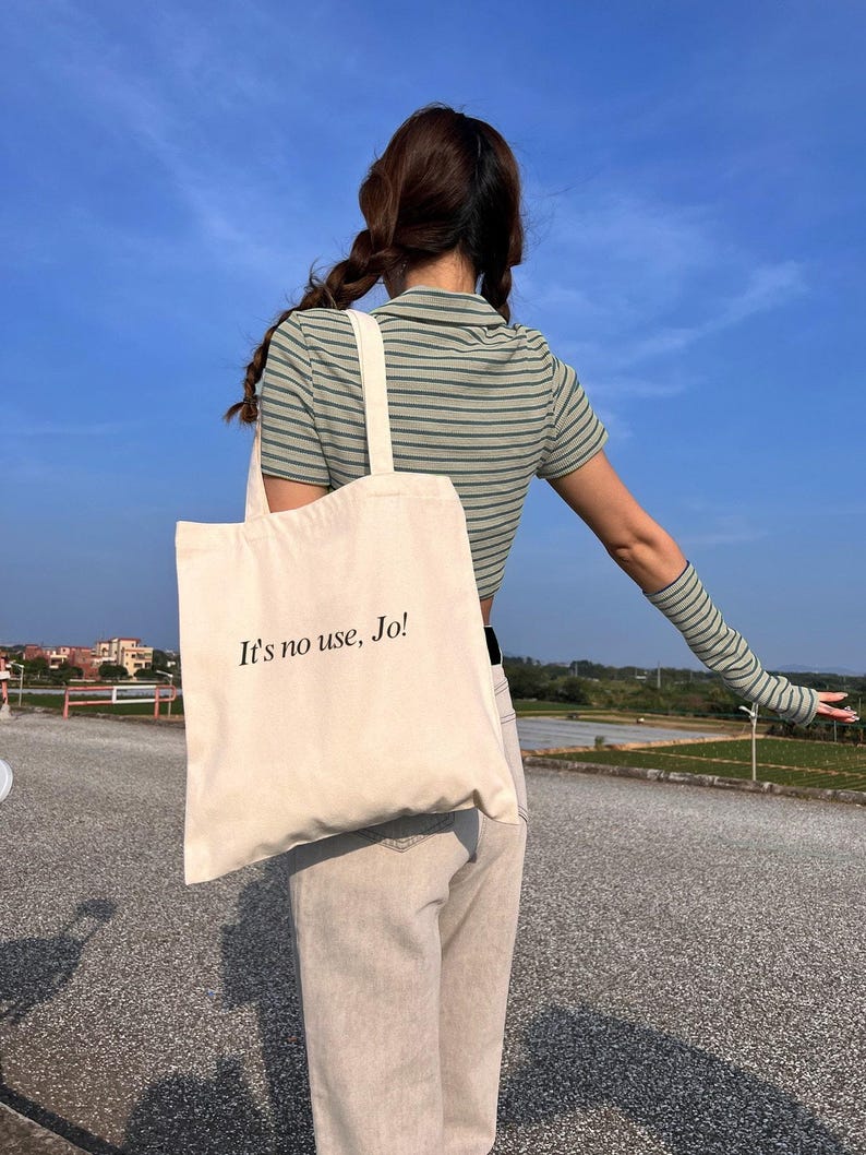 Pode incluir: Uma bolsa tote de lona branca com o texto "It's no use, Jo!" impresso em preto. A bolsa est&aacute; sendo carregada por uma pessoa vestindo uma camisa listrada e jeans.