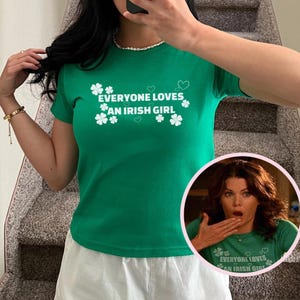 May include: A green t-shirt with the text "EVERYONE LOVES AN IRISH GIRL" in white, along with shamrock and heart graphics. The image also includes a small inset photo of a woman wearing the same shirt.