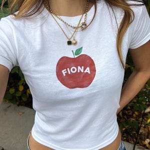 以下が含まれることがあります： 赤いリンゴのグラフィックと「Fiona」という名前がプリントされた白いTシャツ。長い茶色の髪と金のネックレスをした人が着ています。