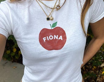 May include: White t-shirt with a red apple graphic and the name "Fiona" printed on it. The shirt is worn by a person with long brown hair and gold necklaces.