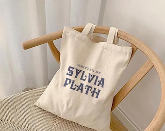 May include: A beige canvas tote bag with the text "Written by Sylvia Plath" printed in blue.