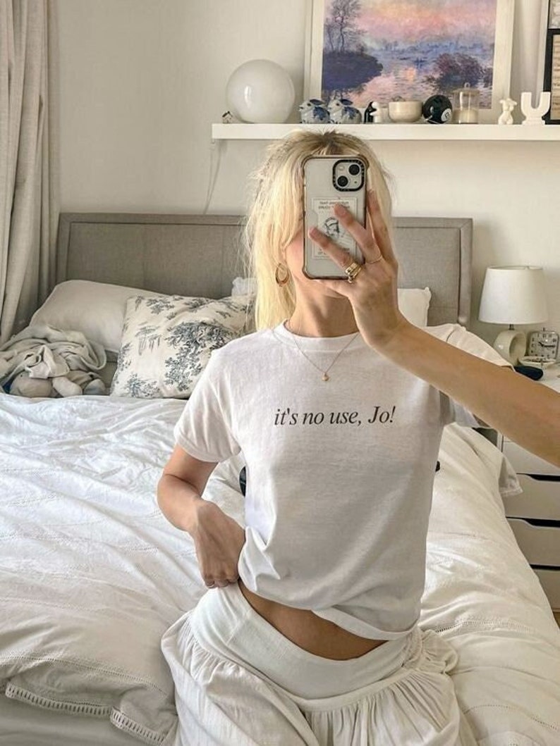 May include: White t-shirt with the text "it's no use, Jo" in black. The shirt is paired with white bottoms. The image is taken indoors, with a bed and a painting in the background.