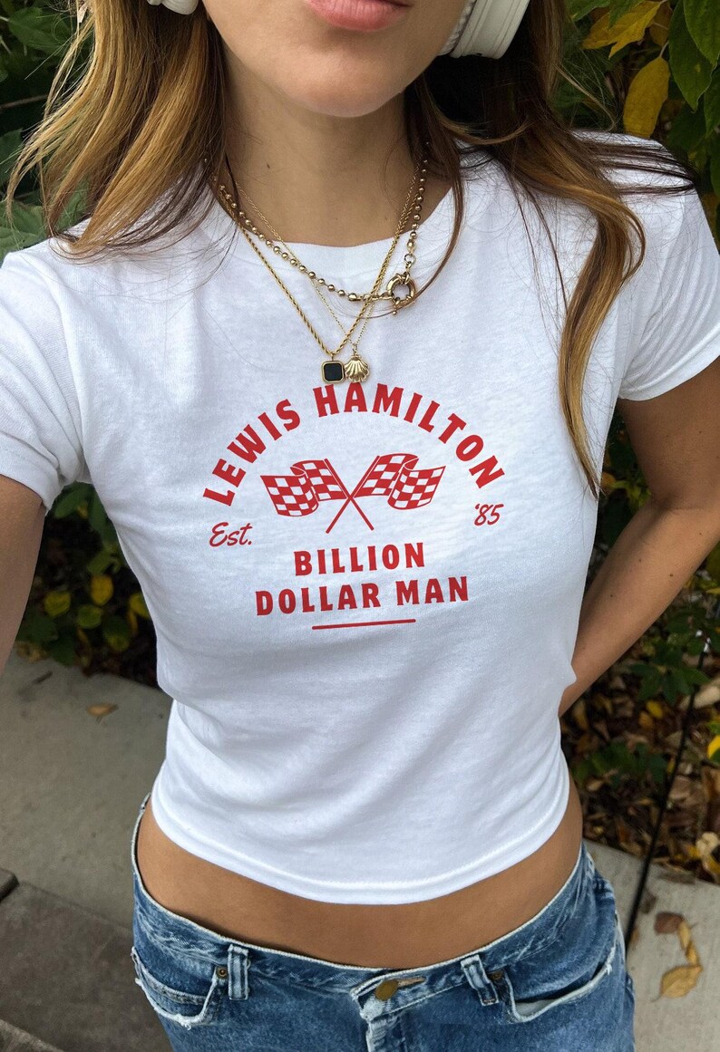 K&ouml;nnte beinhalten: Wei&szlig;es T-Shirt mit einem roten Grafikdruck, auf dem "Lewis Hamilton Est. '85 Billion Dollar Man" mit einem karierten Flaggenmuster steht.