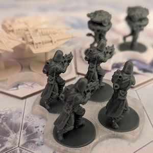May include: Miniature figurines of futuristic soldiers and vehicles, likely for a tabletop game. The soldiers are dark gray, posed with rifles, and stand on circular bases. The vehicles are light beige and have a spaceship-like design. The game board has a hexagonal pattern.