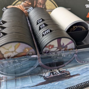 May include: A clear plastic organizer holding stacks of circular game tokens. The tokens are black with colorful illustrations and text. Some tokens have small black tabs with white text. The organizer sits on a game mat.