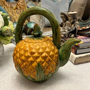 May include: A ceramic teapot shaped like a pineapple with a yellow textured body and green leaves at the base. It features a tall, arched green handle, a green spout, and a matching lid with a pointed finial top, all finished in a glossy glaze.