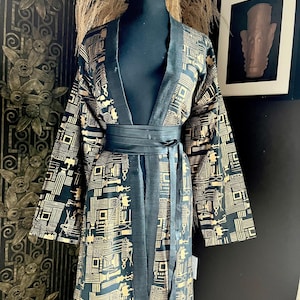May include: A black kimono-style jacket featuring a repeating gold geometric and abstract pattern. The garment includes long, wide sleeves, a solid black trim along the front opening, and a matching black fabric tie belt cinched at the waist.