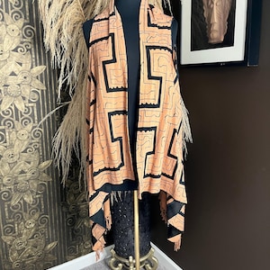May include: A sleeveless vest featuring a light orange base color with a bold black geometric maze-like pattern. The garment has an open front design with a draped silhouette and fringe detailing along the bottom hem, displayed on a mannequin.