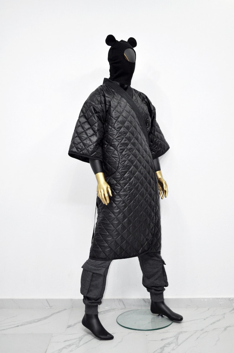 May include: A black quilted robe with a hood and ear flaps. The robe is worn over gray sweatpants with cargo pockets. The model is wearing black socks and black shoes.