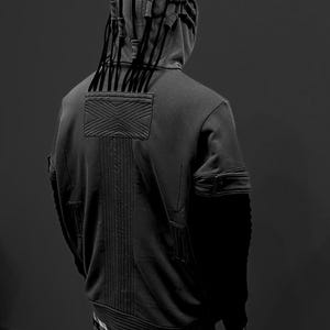 K&ouml;nnte beinhalten: Ein dunkelgrauer Kapuzenpullover mit einem einzigartigen Design, mit einer strukturierten Kapuze mit vertikalen Akzenten und einem strukturierten R&uuml;ckenteil. Der Kapuzenpullover hat eine Tasche an jedem &Auml;rmel und wird mit einem schwarz-wei&szlig; karierten Rock kombiniert.