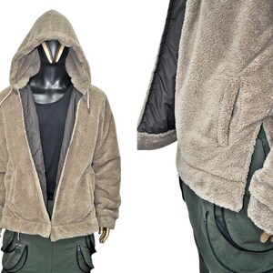 May include: A tan hooded jacket with a full zipper closure. The jacket has a soft, fuzzy texture and a black lining. The jacket is displayed on a mannequin with a black shirt underneath. The jacket has two side pockets and is paired with green cargo pants.