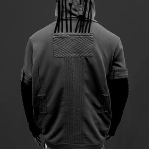 K&ouml;nnte beinhalten: Ein grauer Kapuzenpullover mit einem einzigartigen Design, mit einer Kapuze mit vertikalen schwarzen Akzenten und einem strukturierten Patch auf der R&uuml;ckseite. Der Kapuzenpullover hat schwarze &Auml;rmel und ist mit einem schwarz-wei&szlig;en Karomuster kombiniert.