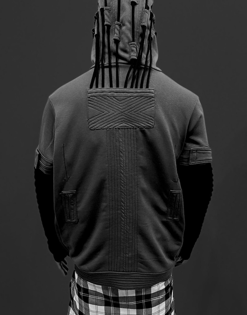K&ouml;nnte beinhalten: Ein grauer Kapuzenpullover mit einem einzigartigen Design, mit einer Kapuze mit vertikalen schwarzen Akzenten und einem strukturierten Patch auf der R&uuml;ckseite. Der Kapuzenpullover hat schwarze &Auml;rmel und ist mit einem schwarz-wei&szlig;en Karomuster kombiniert.