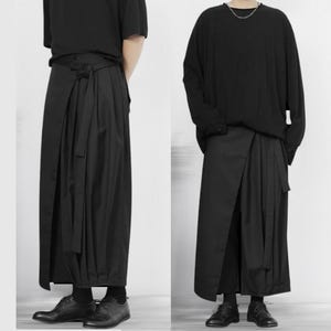 以下が含まれることがあります： 2人のモデルが、長い紐の付いた黒いラップスカートを着用しています。スカートは滑らかで流れるような生地で作られており、ゆったりとしたカジュアルなフィット感です。モデルは黒いシャツと靴を着用しています。