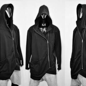 Puede incluir: Chaqueta negra con capucha, cierre de cremallera asim&eacute;trico y bolsillos delanteros. La chaqueta tiene un dise&ntilde;o largo y fluido y se muestra en un maniqu&iacute;. La chaqueta se combina con pantalones grises. La imagen es en blanco y negro.