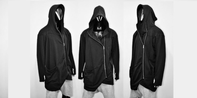 Puede incluir: Chaqueta negra con capucha, cierre de cremallera asim&eacute;trico y bolsillos delanteros. La chaqueta tiene un dise&ntilde;o largo y fluido y se muestra en un maniqu&iacute;. La chaqueta se combina con pantalones grises. La imagen es en blanco y negro.