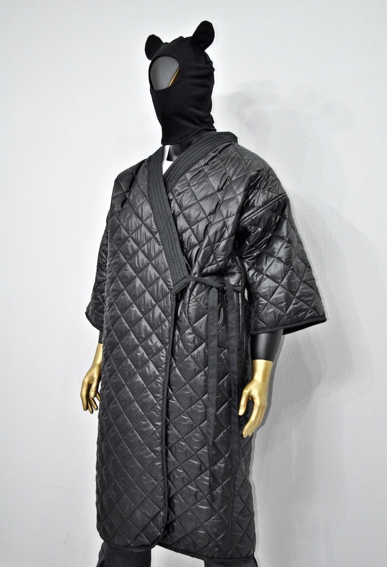 May include: A black quilted robe with a long sleeve and a tie belt. The robe has a diamond pattern and is worn by a mannequin with a black balaclava with bear ears.